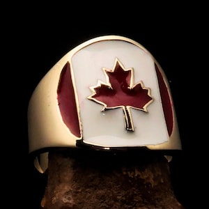 Puede incluir: Un anillo de oro con una hoja de arce roja sobre un fondo blanco. El anillo es una representación de la bandera canadiense.