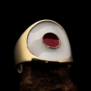 May include: A gold-colored ring with a white enamel oval and a red enamel circle in the center. The ring is set against a dark background, highlighting its polished finish and bold design. This statement ring is a unique piece of jewelry.