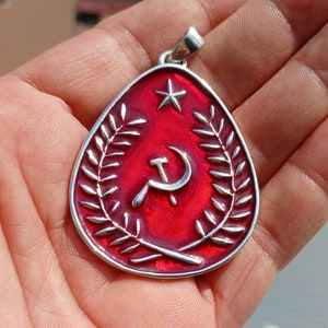 May include: A silver pendant with a red enamel design. The design features a hammer and sickle surrounded by a wreath of leaves. A star is at the top of the pendant.