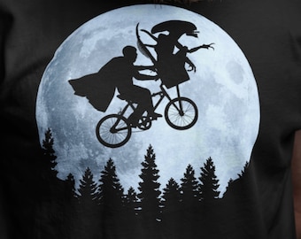 May include: A black t-shirt with a graphic of a silhouette of two figures riding a bicycle in front of a full moon. The figures are riding a bicycle in front of a full moon with trees in the foreground.