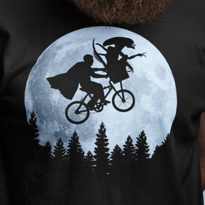 May include: A black t-shirt with a graphic of a silhouette of two figures riding a bicycle in front of a full moon. The figures are riding a bicycle in front of a full moon with trees in the foreground.