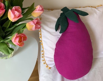 May include: A purple eggplant-shaped pillow with green leaves on a white bed with yellow pom-poms around the edge.
