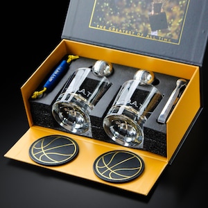 Könnte beinhalten: Eine schwarz-gelbe Geschenkbox mit zwei klaren Whiskeygläsern mit G.O.A.T.-Aufschrift, zwei runden schwarzen Untersetzern mit goldenen Basketball-Linienmustern, einem blauen Kordelzugbeutel und einem silbernen Metallwerkzeug.