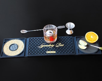 May include: A black rubber bar mat set with the text Legendary Bar and Noblesip, featuring a glass with an ice sphere, a metal mixing spoon, a jigger, a small knife, orange slices, and a circular section filled with brown sugar crystals.