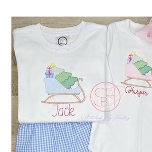 May include: Two white shirts with embroidered Christmas trees and sleds. The shirts are personalized with the names "Jack" and "Harper".