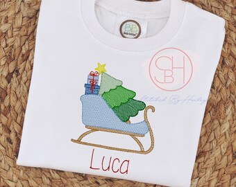 May include: A white t-shirt featuring an embroidered design of a light blue sleigh carrying a green Christmas tree and two small gift boxes topped with a yellow star. The name Luca is embroidered in red below the sleigh graphic.