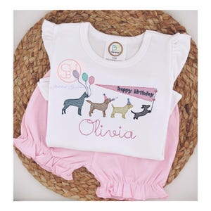May include: A white short-sleeved shirt with a design of four dogs wearing party hats and holding balloons. The shirt has the text "Happy Birthday" and a personalized name "Olivia".