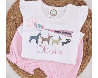 May include: A white short-sleeved shirt featuring a design of four dogs wearing party hats and holding balloons. The shirt has the text "Happy Birthday" and a personalised name "Olivia".