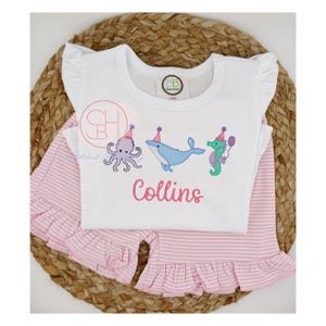 May include: A white, ruffled-sleeve top and matching pink striped shorts set. The top features embroidered sea creatures wearing party hats and the name "Collins" in pink. The set is displayed on a woven mat.