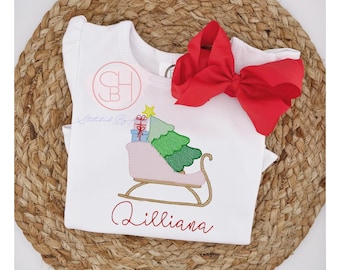 May include: A white short-sleeved shirt featuring an embroidered pink sleigh carrying a green Christmas tree and gifts, topped with a yellow star. The name Lilliana is embroidered in red script below the design, and a large red hair bow is attached.