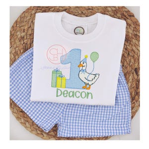 May include: A white short-sleeved shirt with a blue and green embroidered design of a duck with a party hat and presents. The shirt has the number "1" and the name "Deacon" embroidered on it. The shirt is paired with blue and white gingham shorts.