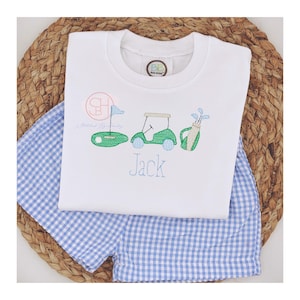 May include: White short-sleeved shirt with a blue and green embroidered golf cart, golf clubs, and a golf ball. The shirt also has a monogram and the name "Jack" embroidered on it.