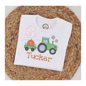 May include: White t-shirt with an embroidered green tractor pulling a wagon with an orange pumpkin. The name "Tucker" is embroidered in orange below the tractor. The shirt is displayed on a woven, brown surface.