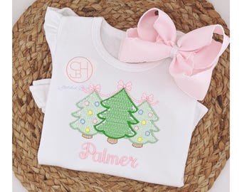 May include: White sleeveless shirt with pink bows and embroidered green Christmas trees with pink and yellow ornaments. The shirt is personalized with the name "Palmer".