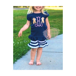 May include: A young girl wearing a navy blue short-sleeved shirt with a pink and white monogram and two embroidered dogs. She is also wearing white and navy blue striped shorts with a ruffle trim.