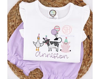 May include: A white short-sleeved top with a ruffled neckline and a purple pair of shorts with ruffles around the legs. The top has an embroidered design of a goose, a cow, a pig, and balloons. The text "Stitched by Hubby" and "Anniston" are also embroidered on the top.