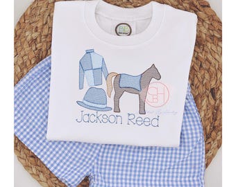Puede incluir: Conjunto de niño pequeño de color blanco bordado con un diseño de jockey, caballo y sombrero. El conjunto incluye un pantalón corto de cuadros azules y blancos y una camisa de manga larga blanca con el nombre "Jackson Reed" bordado.