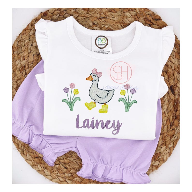 May include: A white short-sleeved shirt with a ruffle trim on the sleeves. The shirt features an embroidered design of a goose with a pink bow, surrounded by yellow and purple flowers. The name "Lainey" is embroidered below the goose. The shirt is paired with a lavender pair of shorts with a ruffle trim.