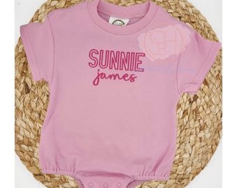 May include: A light pink baby onesie with short sleeves and a round neckline. The text "SUNNIE james" is printed in a darker pink, stacked vertically. The onesie is displayed on a woven, circular placemat.