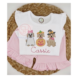 May include: A white Halloween-themed outfit with a top and bottoms. The white top features three embroidered dogs dressed as a ghost, a pumpkin, and a witch. The name "Cassie" is embroidered in pink. The bottoms are pink and white striped with a ruffled hem.