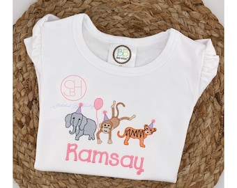 Pode incluir: Camiseta branca de manga curta com um elefante, um macaco e um tigre bordados em rosa usando chapéus de festa. A camiseta também tem o nome "Ramsay" bordado em rosa.