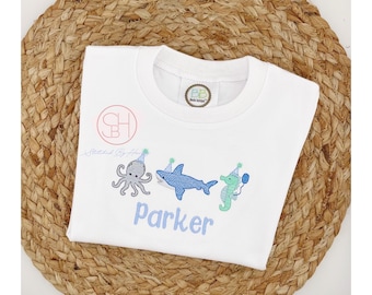 May include: White t-shirt with an embroidered design of an octopus, shark, and seahorse wearing party hats. The text "Parker" is embroidered below the design.