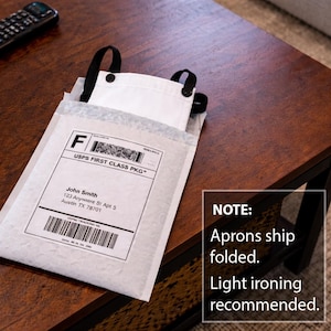Pu&ograve; includere: Un grembiule bianco con spalline nere &egrave; parzialmente visibile all'interno di una busta a bolle bianca. La busta ha un'etichetta di spedizione con la scritta "USPS FIRST CLASS PKG" e l'indirizzo del destinatario. Il grembiule &egrave; piegato. Si consiglia una stiratura leggera.