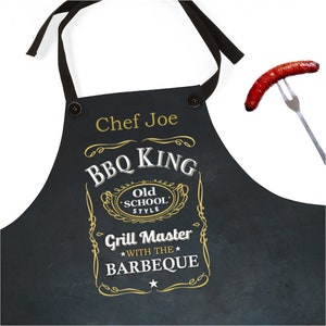 Puede incluir: Delantal negro con un dise&ntilde;o dorado y blanco que dice "BBQ King Old School Style Grill Master With The Barbeque". El delantal tiene una correa negra y un bot&oacute;n en la parte superior. El texto "Chef Joe" est&aacute; impreso en el delantal.