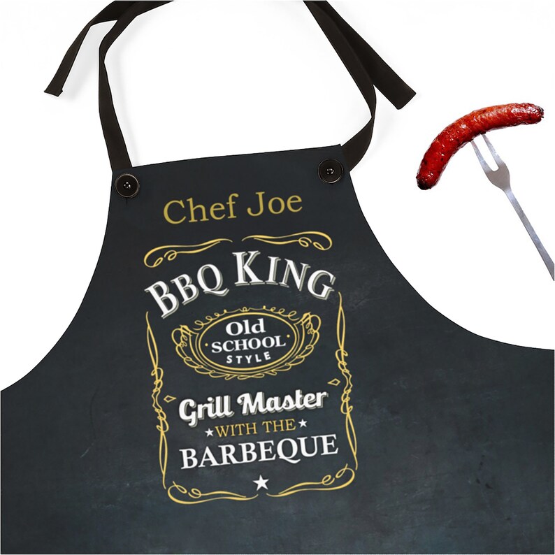 Puede incluir: Delantal negro con un dise&ntilde;o dorado y blanco que dice "BBQ King Old School Style Grill Master With The Barbeque". El delantal tiene una correa negra y un bot&oacute;n en la parte superior. El texto "Chef Joe" est&aacute; impreso en el delantal.