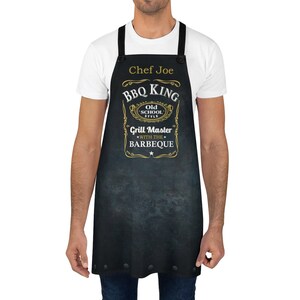 Puede incluir: Delantal negro con un dise&ntilde;o dorado que dice "BBQ King Old School Style Grill Master with the Barbeque". El delantal tiene una etiqueta de nombre que dice "Chef Joe".