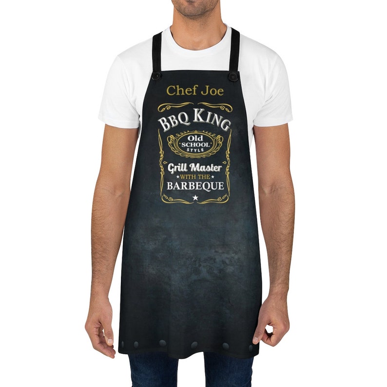 Puede incluir: Delantal negro con un dise&ntilde;o dorado que dice "BBQ King Old School Style Grill Master with the Barbeque". El delantal tiene una etiqueta de nombre que dice "Chef Joe".