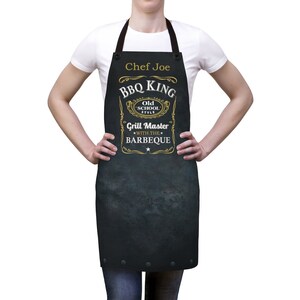 Puede incluir: Delantal negro con un dise&ntilde;o dorado que dice "BBQ King Old School Style Grill Master With The Barbeque" y "Chef Joe" en la parte superior.