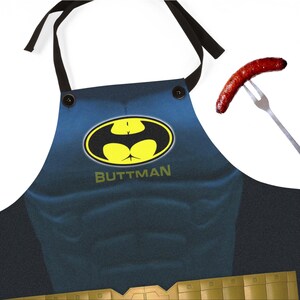 May include: A blue apron with a yellow and black emblem that says "BUTTMAN". The apron has black straps and buttons. A cooked sausage is impaled on a fork to the right of the apron.