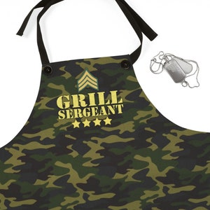 May include: Camouflage-patterned apron with the text "GRILL SERGEANT" in gold lettering, along with four gold stars and a military rank insignia. The apron has black ties and buttons. A pair of dog tags are also visible.
