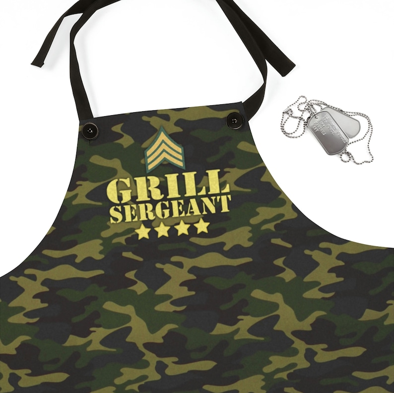 May include: Camouflage-patterned apron with the text "GRILL SERGEANT" in gold lettering, along with four gold stars and a military rank insignia. The apron has black ties and buttons. A pair of dog tags are also visible.