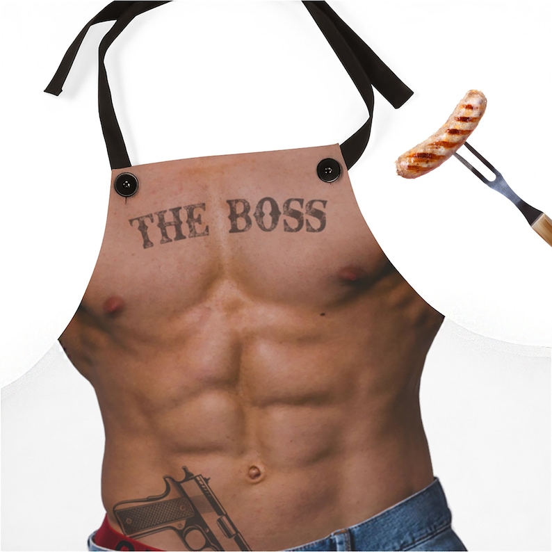 Pu&ograve; includere: Grembiule bianco con una grafica di un busto muscoloso, la scritta "THE BOSS" stampata sul petto. Il grembiule ha lacci e bottoni neri. Una salsiccia grigliata &egrave; tenuta da una forchetta nell'immagine.