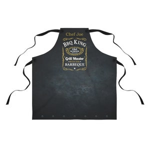 Puede incluir: Delantal negro con un dise&ntilde;o dorado que dice "BBQ King Old School Style Grill Master with the Barbeque". El delantal tiene una etiqueta de nombre que dice "Chef Joe".