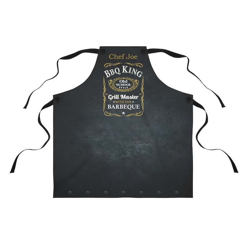 Puede incluir: Delantal negro con un dise&ntilde;o dorado que dice "BBQ King Old School Style Grill Master with the Barbeque". El delantal tiene una etiqueta de nombre que dice "Chef Joe".