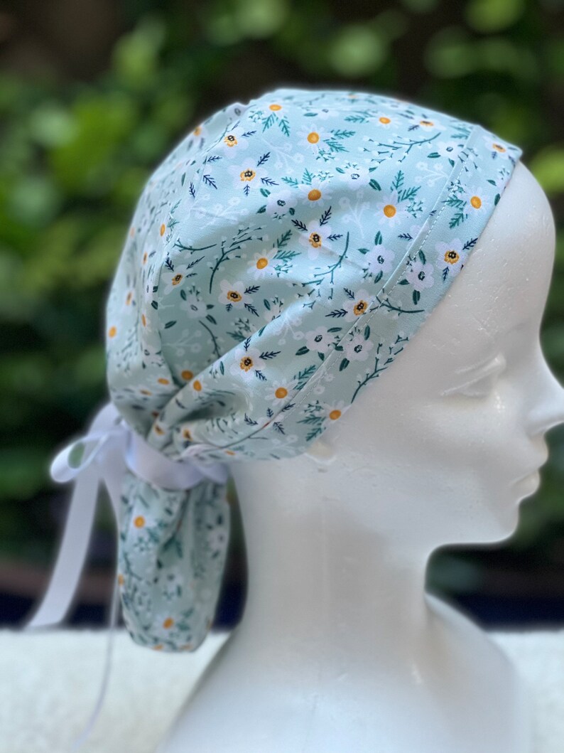 K&ouml;nnte beinhalten: Eine hellblaue OP-M&uuml;tze mit wei&szlig;em Blumenmuster. Die M&uuml;tze hat einen Bindfaden am R&uuml;cken.