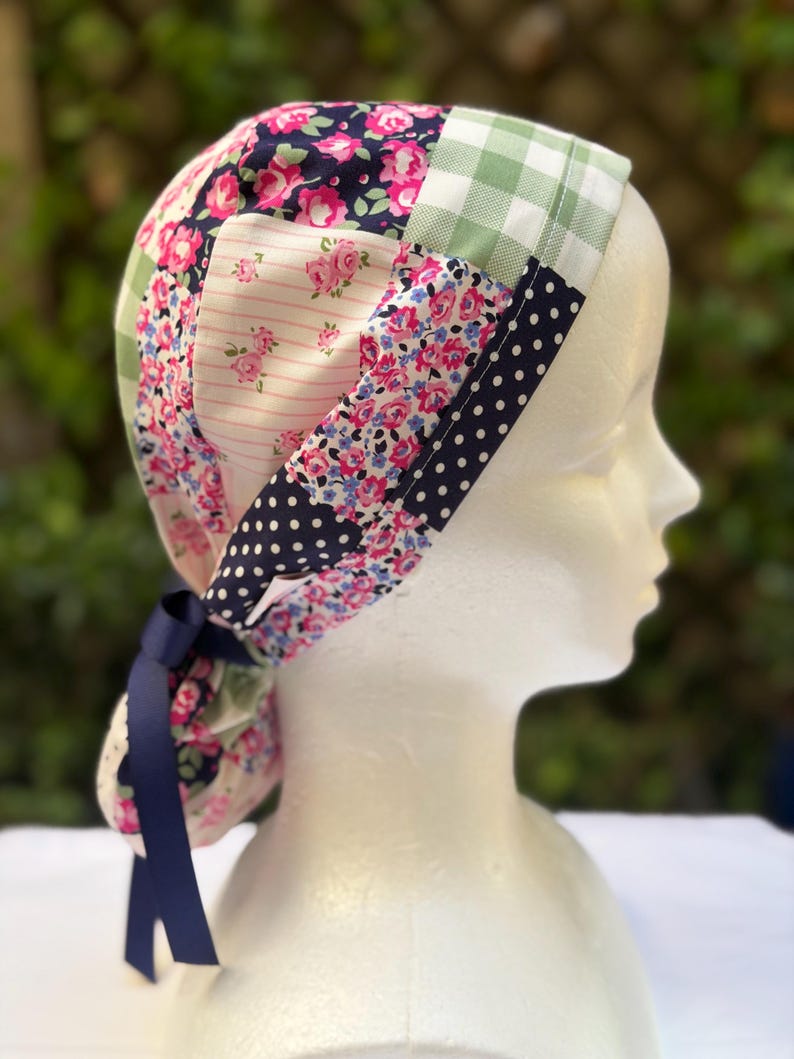 May include: A patchwork scrub cap featuring a variety of floral and polka dot patterns in pink, blue, and white. The cap includes a navy blue ribbon for tying. The cap is displayed on a white mannequin head.
