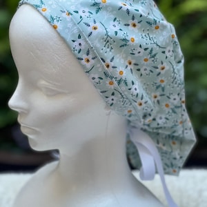 K&ouml;nnte beinhalten: Eine hellblaue OP-M&uuml;tze mit einem wei&szlig;en und gelben Blumenmuster. Die M&uuml;tze hat einen wei&szlig;en Band hinten.