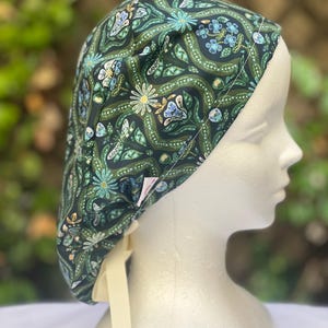 May include: A dark green patterned scrub cap featuring a floral design with white, blue, and yellow accents. The cap has a cream-colored tie and is displayed on a mannequin head. The fabric has a repeating pattern of flowers and leaves.