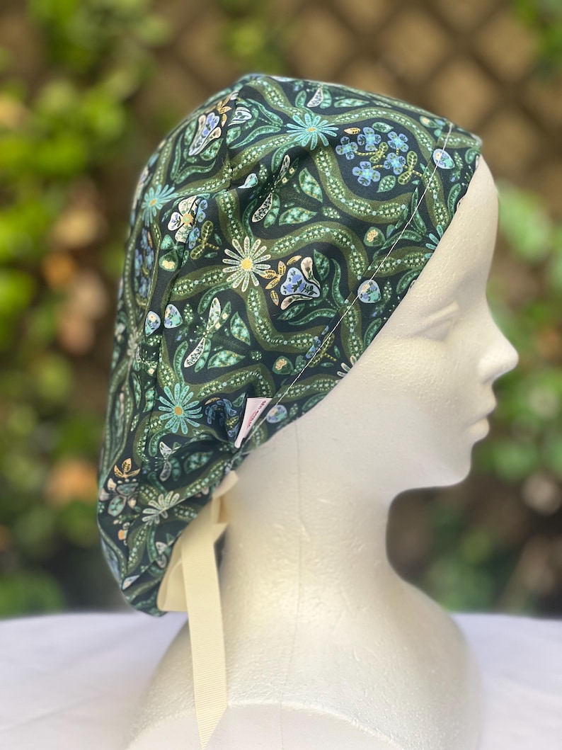 May include: A dark green patterned scrub cap featuring a floral design with white, blue, and yellow accents. The cap has a cream-colored tie and is displayed on a mannequin head. The fabric has a repeating pattern of flowers and leaves.