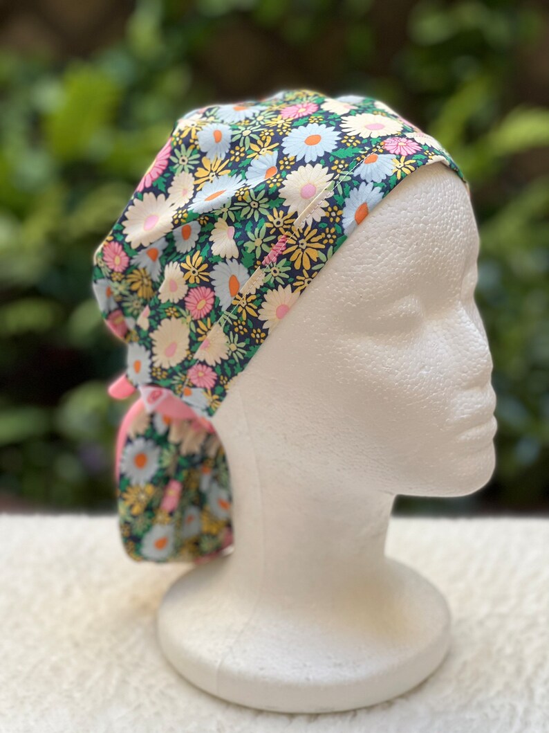 May include: A floral print surgical scrub hat with a pink tie. The hat has a dark blue background with white, pink, and yellow flowers.
