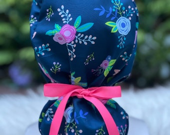 May include: A dark blue scrub hat with a floral print in shades of pink, blue, green, and white. The hat has a pink ribbon tied at the back.