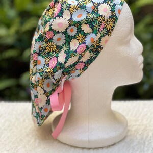 May include: A black scrub cap with a floral print featuring pink, yellow, and white daisies. The cap has a pink ribbon tie.