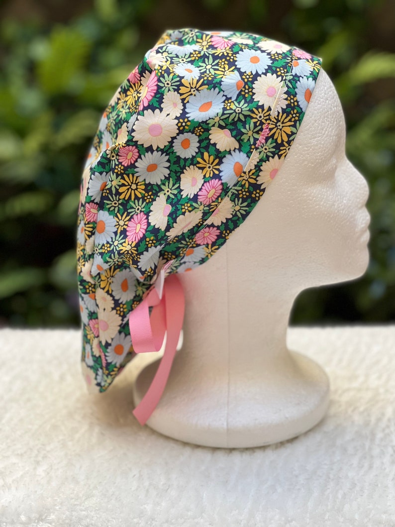 May include: A black scrub cap with a floral print featuring pink, yellow, and white daisies. The cap has a pink ribbon tie.