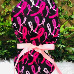 May include: A black scrub hat with a pink ribbon pattern. The hat is tied with a pink ribbon.