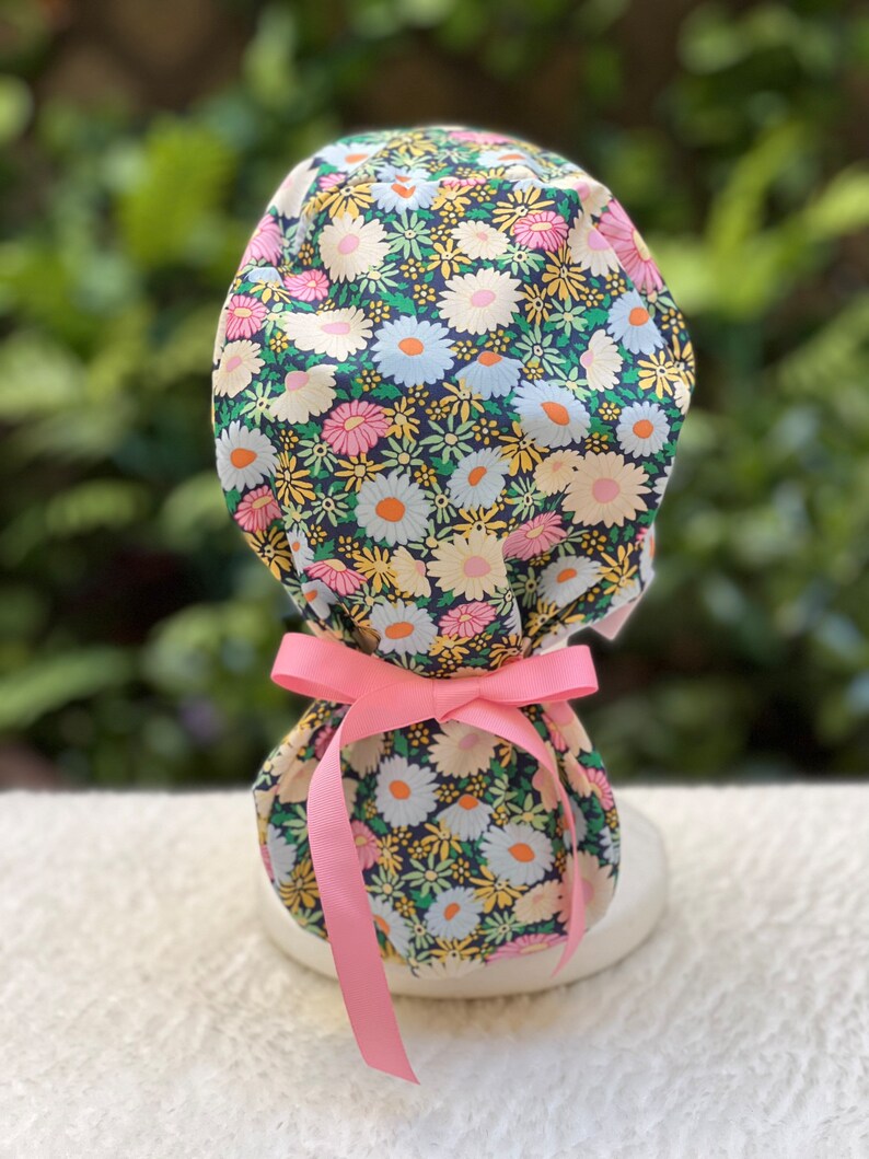 May include: A surgical cap with a floral print featuring pink, yellow, and white flowers on a dark blue background. The cap has a pink ribbon tie.