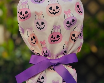May include: A white scrub cap patterned with pink and purple jack-o'-lanterns with black faces, tied with a large purple bow. The cap is ideal for Halloween or adding a festive touch. The cap's design is perfect for the spooky season.